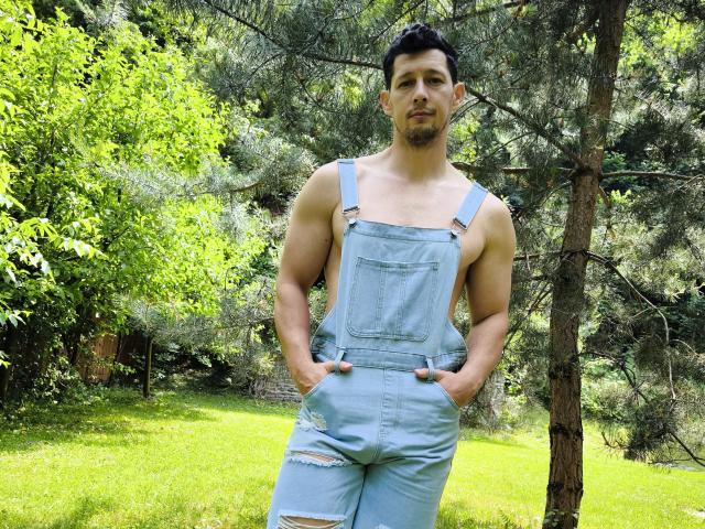 BrutusK mit muskulösen Armen, der leichte, zerrissene Jeans-Overalls in Hellblau trägt, steht in einer grasbewachsenen, bewaldeten Gegend, die Hände in den Taschen, und blickt mit ernstem Gesichtsausdruck in die Kamera..