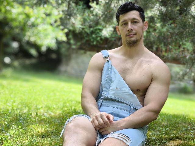 BrutusK, shirtless with muscular arms, wearing blue denim overalls, sitting on grass, hands on knees, short dark hair, slight beard, looking at camera, green background, sunny day.
