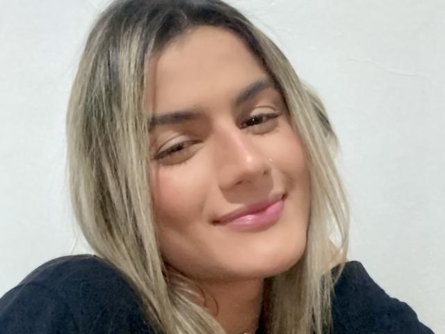 BabeZina, com cabelos loiros e levemente ondulados, sorrindo suavemente, vestindo uma blusa preta, tem uma fenda charmosa no nariz e uma maquiagem leve. Seus lábios rosados estão ligeiramente entreabertos..
