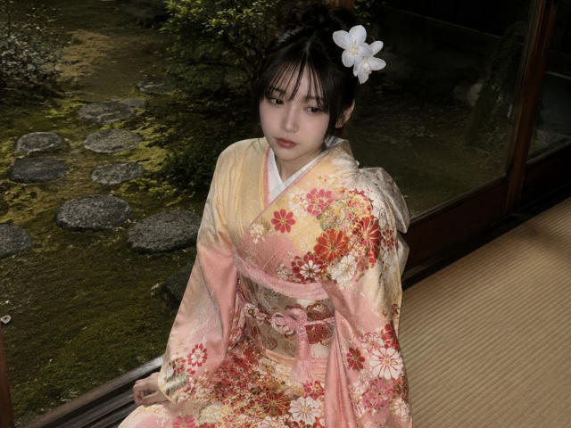 Annetta Hatchcock, com cabelo escuro, uma flor branca no cabelo, vestindo um quimono floral rosa e vermelho, sentada em um ambiente interno, olhando para a câmera, com uma expressão suave, em estilo tradicional japonês..