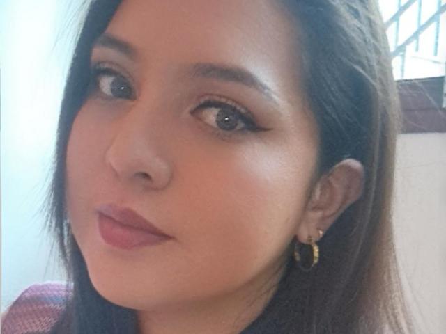 Paula tem a pele bronzeada, olhos castanhos e cabelos longos castanhos, usa brincos de argola dourados e batom rosa, com um leve sorriso no rosto..