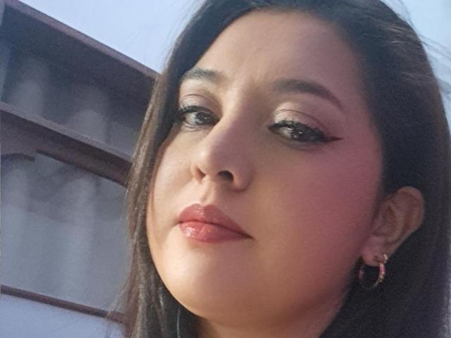 PaulaAnd's face, showing glossy pink lips, subtle eyeliner, and brown hair, with hoop earrings, looking slightly to the side. Her skin is light with a slight blush.