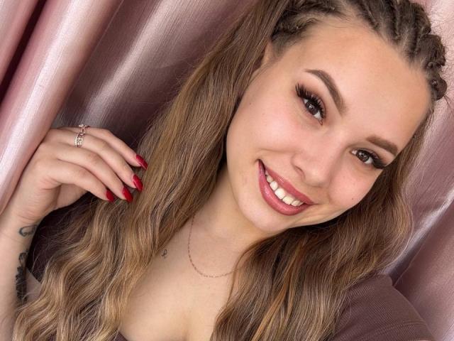 Niferttiti con lunghi capelli castano ondulati raccolti in trecce, unghie rosse, che sorride mostrando i denti bianchi, rossetto rosa, indossa una collana, un anello e una maglia trasparente, con un tatuaggio carino..