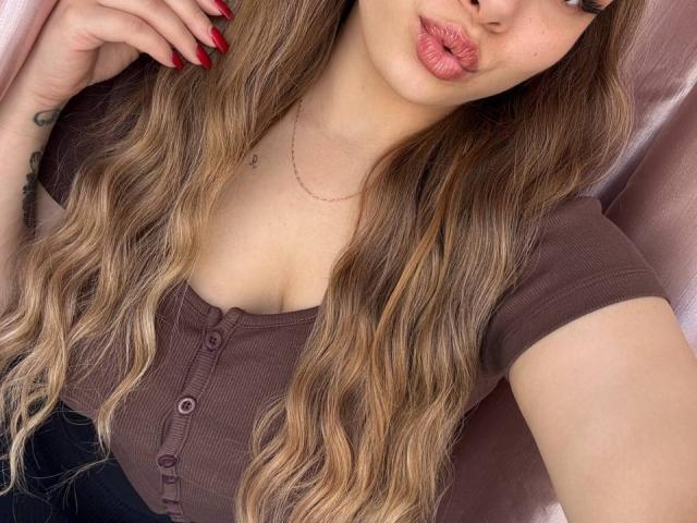 Niferttiti, avec de longs cheveux blonds ondulés, des ongles rouges, un haut marron à boutons, laissant entrevoir ses petites seins, des lèvres roses et pulpeuses, un tatouage au poignet, allongée, avec un rideau rose en arrière-plan..
