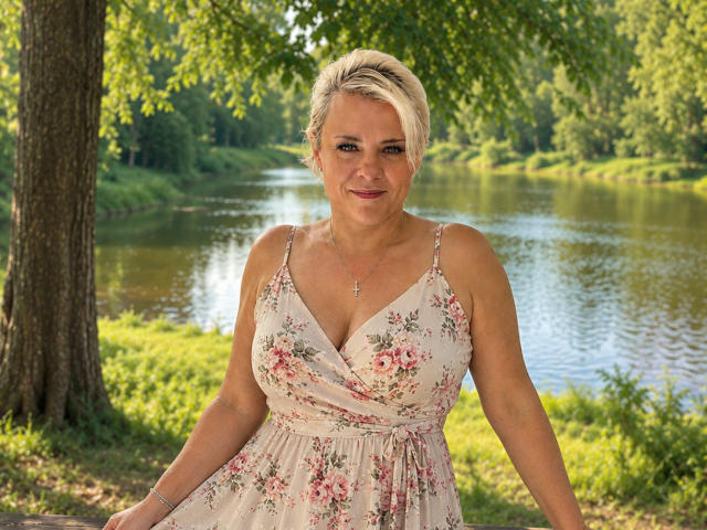 CrystalVibe has short blonde hair, floral dress with pink roses, medium tits, smiling, standing by a tree, near a pond, wearing a necklace, sunlight, green background.