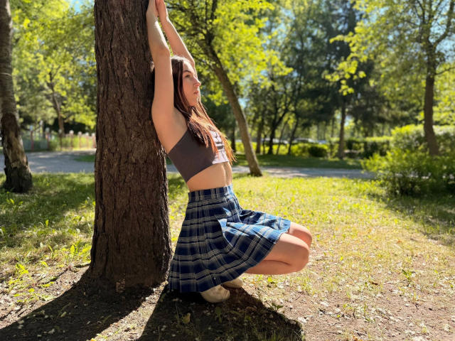 SweetCookies: squat fa una serie di esercizi con i glutei contro un albero in un parco illuminato dal sole, con le braccia alzate, indossa un reggiseno sportivo marrone e una gonna a quadri blu, i glutei e i seni sono leggermente visibili, lunghi capelli castani..