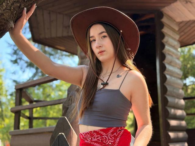 SweetCookies, con lunghi capelli castani, indossa un cappello marrone, un top corto grigio, una bandana rossa e una collana, ha piccoli seni e un tatuaggio a forma di cuore sul petto, ed è in piedi vicino a un albero..