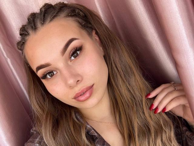 Niferttiti hat eine hellbraune Haut, Zöpfe, dicke Augenbrauen, lange Wimpern, rosafarbenen Lippenstift, rote Nägel und ein süßes Gesicht. Sie trägt eine karierte Bluse mit einem pinken Vorhang..