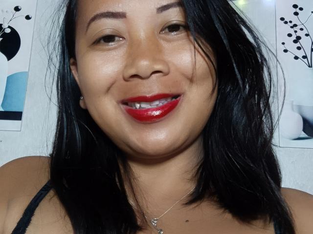SarahFontaineX tiene una piel marrón media, cabello negro de largo medio, un lápiz labial rojo brillante y una ligera sonrisa, y lleva un top negro con tirantes finos y un collar pequeño..