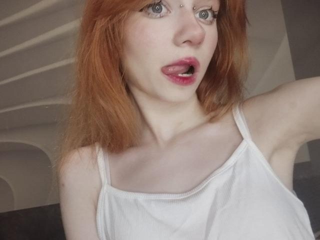 JamieLathrum with red hair, white tank top, and pierced nose, mouth open, showing pink lips, blue eyes, and fair skin, raising an arm.