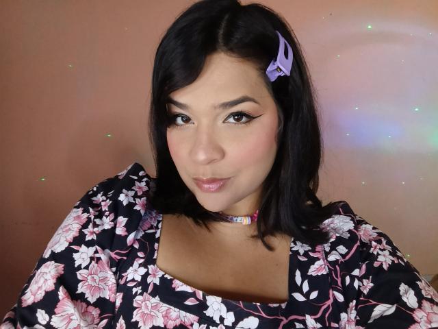 AntonelaPark has black hair with a purple hair clip, wearing a floral dress, and a colorful choker, with winged eyeliner and pink lipstick, looking at the camera.