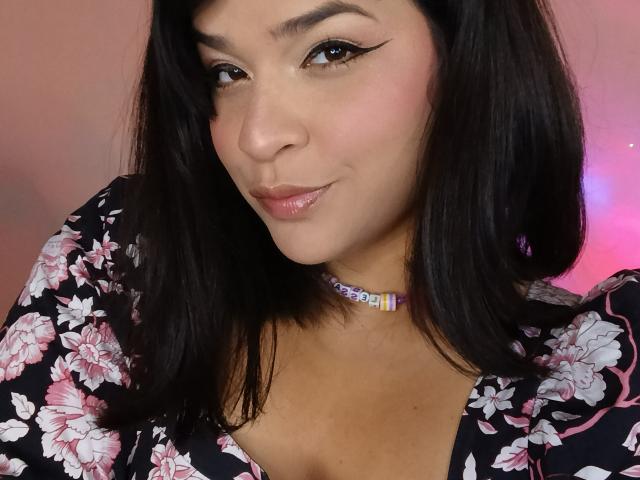 AntonelaPark with dark hair, wearing a floral dress, and a colorful choker, has pouty lips, winged eyeliner, and a slight smile, showing her tits in the photo.