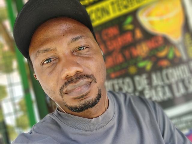 Anaconda69cm is a black man with a beard and mustache, wearing a black cap and gray shirt, smiling slightly with green eyes, and a slightly sweaty forehead.
