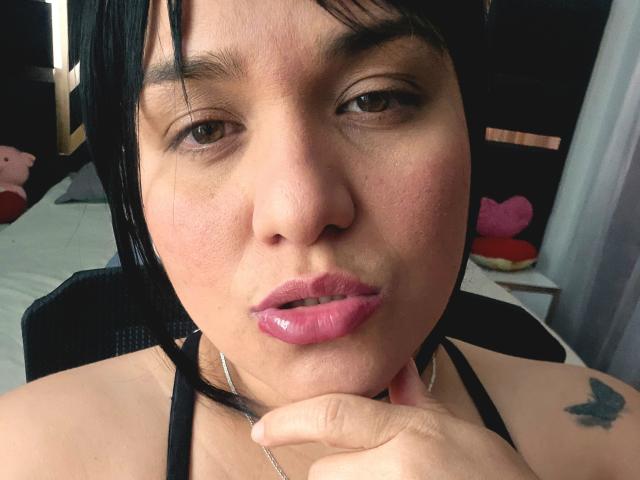 Viviana Thompson tem lábios rosados e brilhantes, cabelo preto e uma tatuagem de borboleta no ombro direito. Ela está tocando o queixo com um colar de prata. Seus olhos castanhos estão fixos para frente..