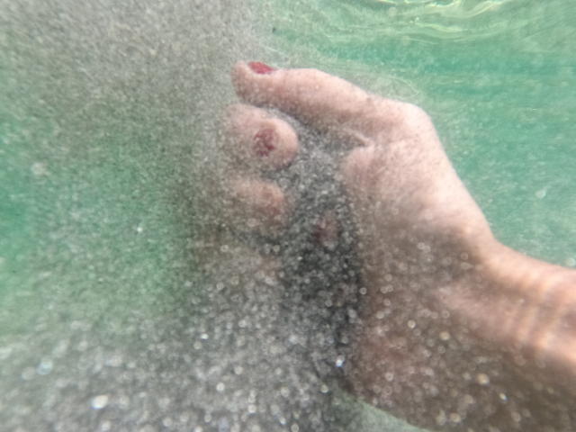 De hand van OrianaLaFrancaise, met rood gelakte nagels, drukte tegen een helder, borrelend oppervlak onder water, waarbij de vingers lichtjes wazig waren en het water een groene tint had..