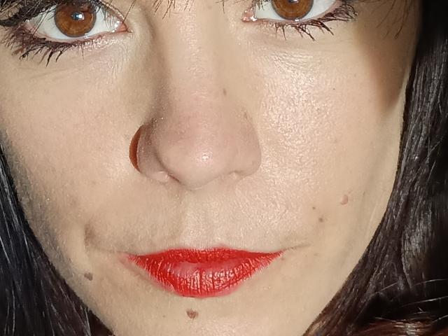 OrianaLaFrancaise hat ein Gesicht mit braunen Augen, rotem Lippenstift, kleinen Sommersprossen auf der Haut, schwarzen Haaren, einer leicht glänzenden Nase und sichtbarer Hautstruktur. Ihre Lippen sind glänzend und leicht geöffnet..