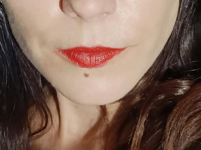 Het gezicht van OrianaLaFrancaise, met bleke huid, rode lippenstift en een klein beauty markje net onder haar onderlip, met bruin haar aan beide kanten..