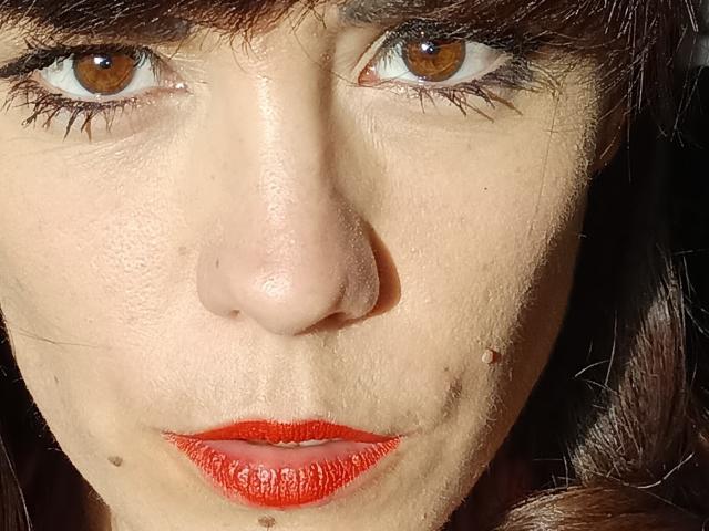 Il viso di OrianaLaFrancaise, occhi marroni, rossetto rosso, naso leggermente pronunciato, pelle chiara con piccole lentiggini, capelli castano scuro, sguardo diretto verso la fotocamera, illuminata dal sole, pelle con texture, espressione intensa..