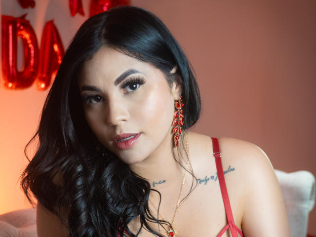 EllieeSmith has black wavy hair, red lipstick, and red earrings. She's wearing a red bra, showing small tattoos on her chest, with her tits slightly visible.
