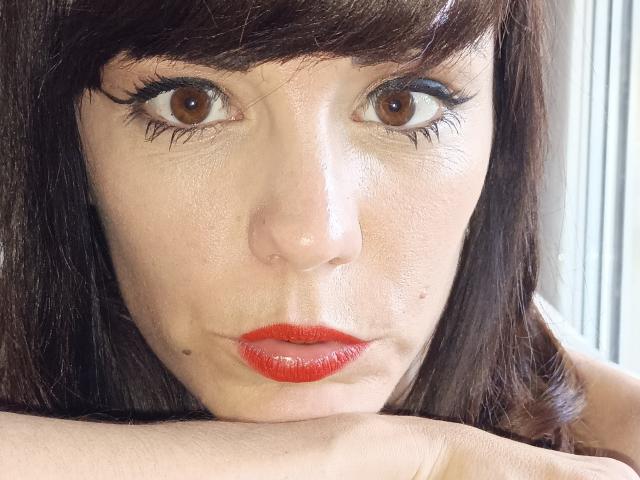 OrianaLaFrancaise a les yeux marron, du rouge à lèvres, et des cheveux brun foncé, ses bras reposant sous son menton, regardant directement l'appareil photo avec une expression légèrement boudeuse..