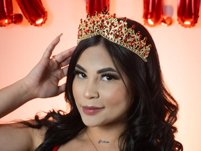 EllieeSmith with long black hair, wearing a gold and red jeweled crown, has her hand near her head, showing a subtle smile and a small nose ring. Boobs visible.