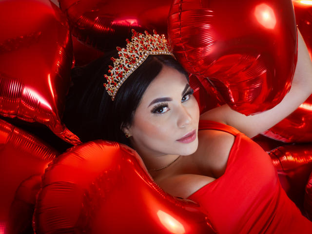 EllieeSmith with red balloons, wearing a sparkly tiara, red dress, and showing her big tits, looking up with makeup and dark hair. Red balloons surround her.