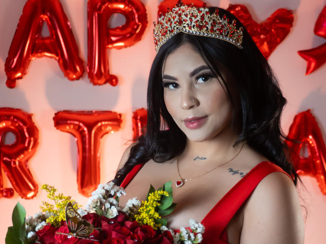 EllieeSmith with black hair, wearing a red dress, tiara, and necklace, holding a bouquet, showing her tits, and a small tattoo on her chest, smiling at the camera.