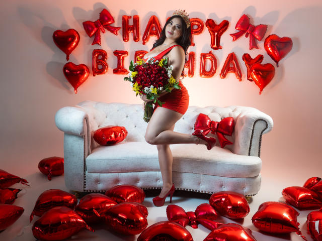 EllieeSmith with red dress, high heels, and crown, holding bouquet, standing on one leg, white tufted couch, red heart balloons, red heart-shaped pillows, and red wall decor.
