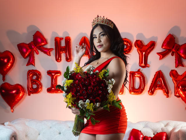EllieeSmith with dark hair, red dress, and gold crown, holding a red rose bouquet, standing in front of red heart and bow balloons, showing off her big boobs.