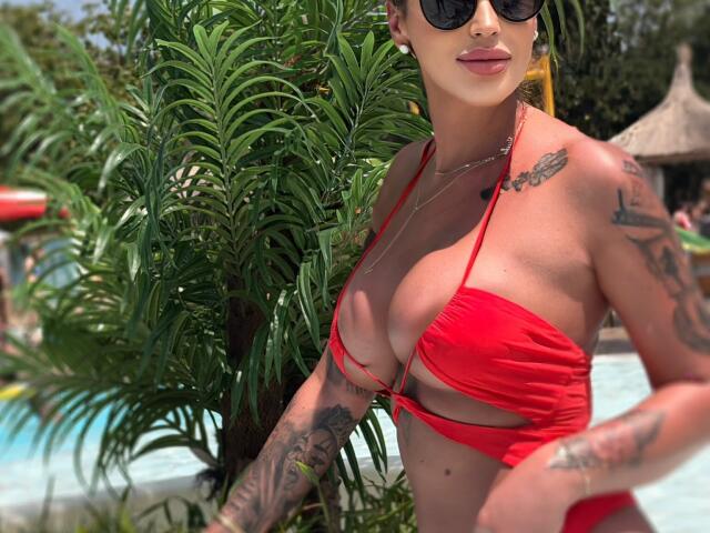 NicolSNaya in a red bikini, showing off her big tits and tattoos, wearing black sunglasses, standing near a palm tree, by a pool. Her lip tattoo is visible.