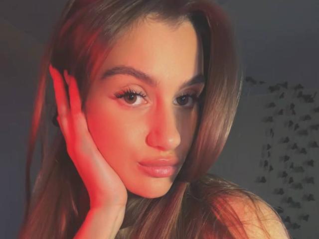 EllenKienbaum with red-lit face, long brown hair, and subtle makeup, resting her hand on her cheek, brown eyes, and glossy pink lips, in a soft-focused selfie.