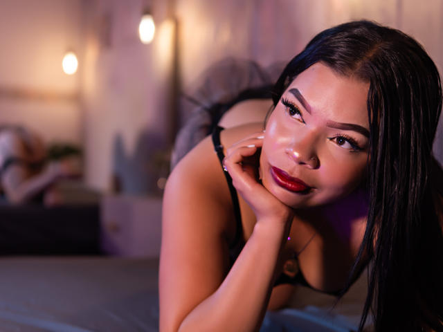 AdrianaPaul, con cabello oscuro, lápiz labial rojo y lencería negra, inclinada hacia adelante con la mano en la mejilla, mostrando sus pechos y glúteos, mirando a la cámara con una sonrisa sensual..
