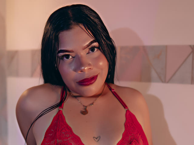 AdrianaPaul with dark wet hair, red lace bra, heart tattoo below tits, red lipstick, black eyeliner, gold necklace, smiling at camera. Warm lighting, slight shadow on face.