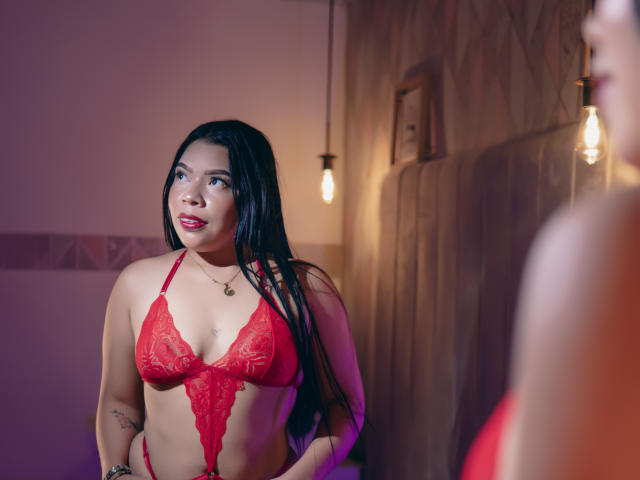 AdrianaPaul met lang zwart haar, een rode kantbeha, waarbij haar borsten zichtbaar zijn, die in een spiegel kijkt, rode lippenstift, een tatoeage op haar arm, een hangerketting, warm licht, een houten muur..