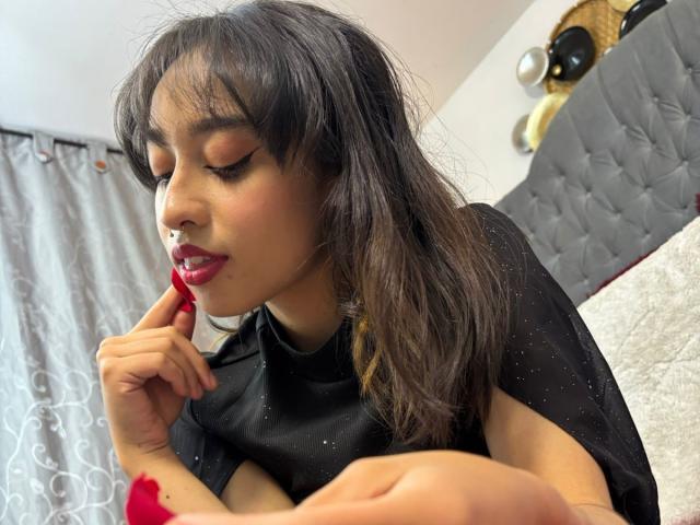 CamiVargas with dark hair, wearing a black top, red lipstick, and sparkly eyeshadow, touching her lip with a red-manicured finger, in a slightly messy room.