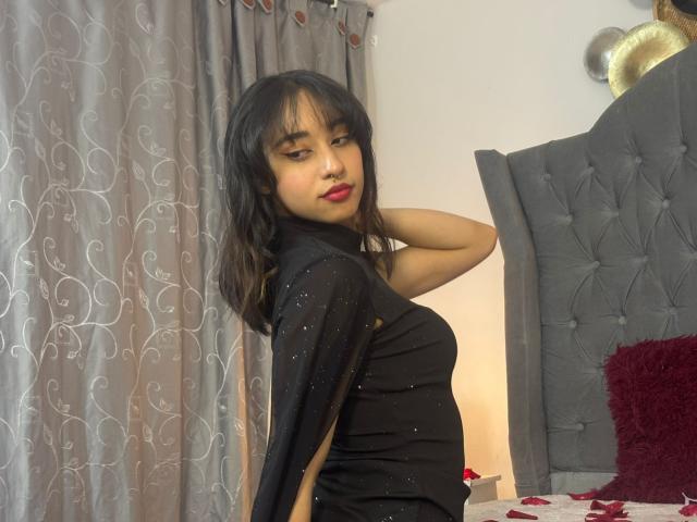 CamiVargas with long black hair, wearing a black sheer dress with sparkles, red lipstick, standing by a grey tufted headboard, hand behind head, looking over shoulder.