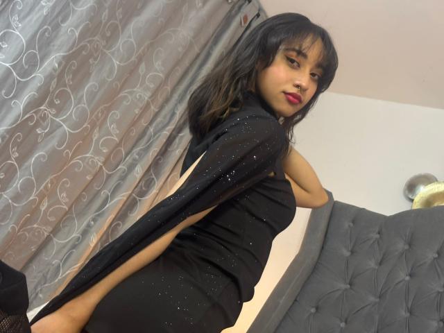 CamiVargas with black hair, red lipstick, wearing a black sparkly dress, showing her ass, looking over her shoulder, standing in front of a grey tufted headboard.
