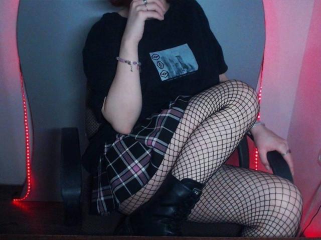 AyumiBabe, les jambes croisées, portant des bas résille, des bottes noires, une jupe à carreaux, un t-shirt noir et un bracelet violet, assise sur une chaise avec des lumières LED rouges..