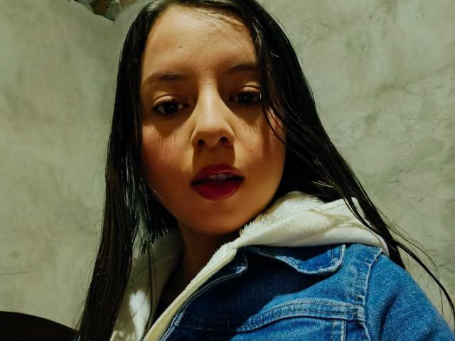 VioletaNova with wet black hair, wearing a blue denim jacket with a white hoodie, has red lipstick and a slightly open mouth, looking at the camera.