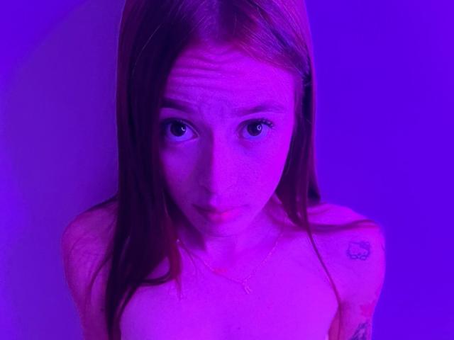 KittyMiller with red hair, purple-lit, looking up with a slightly confused expression, small tattoo on right shoulder, topless, visible collarbone, and a subtle necklace.