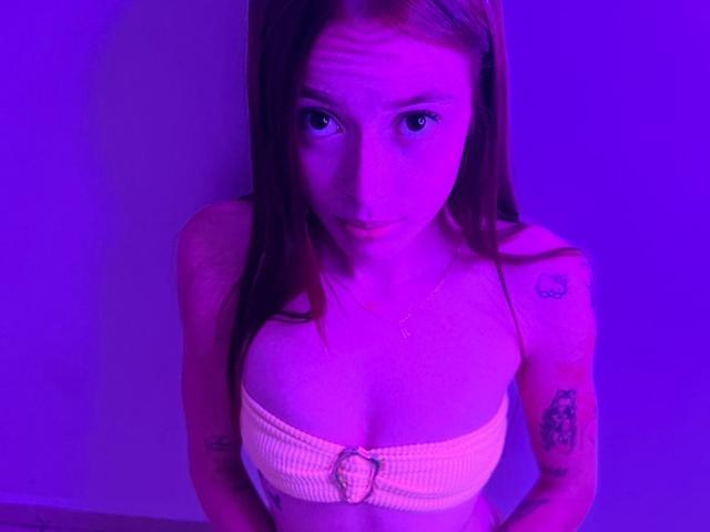 KittyMiller with pinkish-purple lighting, wearing a strapless yellow tube top, showing her small tits and tattoos on her arms, looking up with a slight pout.