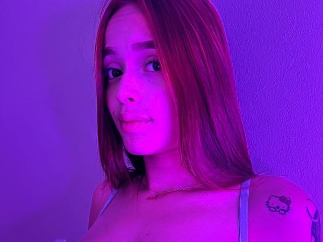 KittyMiller met roze-paarse verlichting, lang rood haar, kleine borsten, een tatoeage op de rechter schouder, een subtiele glimlach, gekleed in een top met dunne bandjes en een gouden ketting..