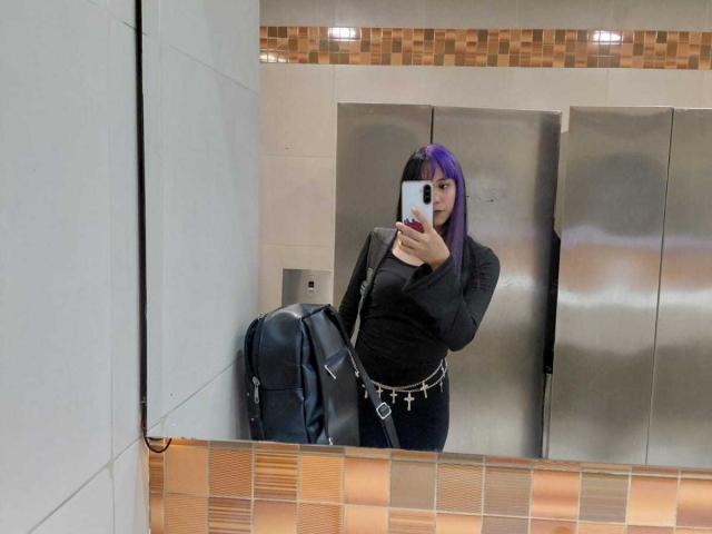 RaveenMoonn, avec des cheveux violets, un t-shirt noir, un pantalon noir, une ceinture avec des clés, tenant un téléphone, prenant un selfie devant un miroir, debout devant des portes d'ascenseur en métal, un sac à dos noir sur la gauche..