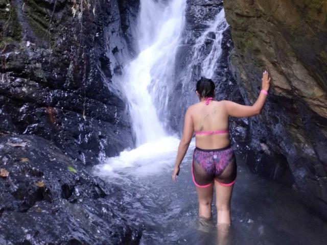 Novarose stands in a rocky waterfall, wearing a pink bikini top and see-through pink and black lace shorts, with her back to the camera, holding a rock.