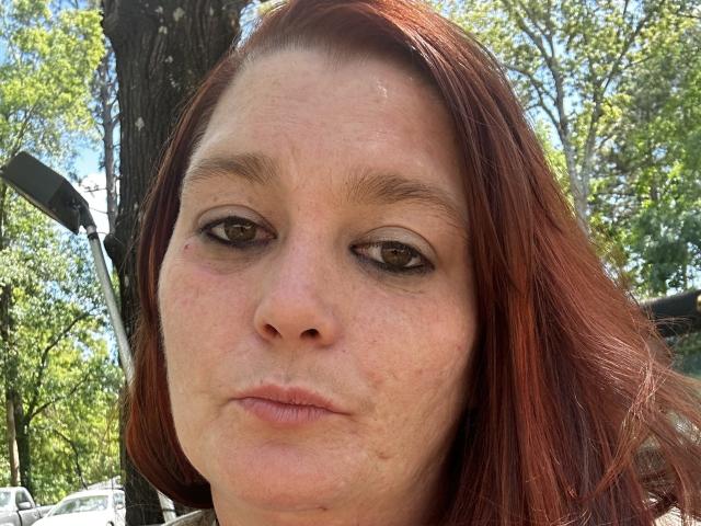 Ashlleyy69 tem cabelo ruivo, olhos castanhos e um rosto com lábios finos e sardas, com uma expressão ligeiramente entediada ou irritada, estando ao ar livre com uma árvore e uma bicicleta ao fundo..