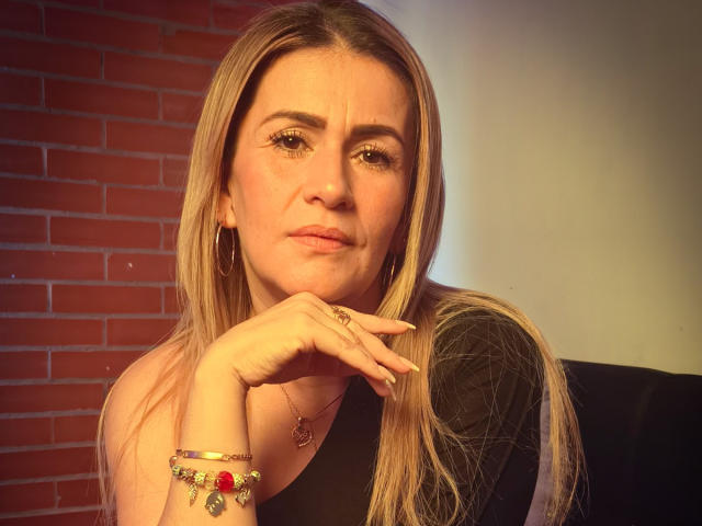 CamilleMullerr heeft lang blond haar, draagt een zwarte top, oorringen en kleurrijke armbanden. Ze leunt haar kin op haar hand en kijkt lichtelijk geïrriteerd, met een subtiele frons..