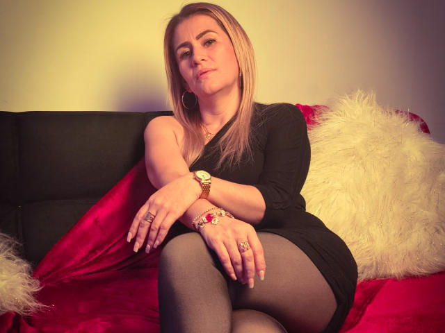 CamilleMullerr sitzt auf einem schwarzen Sofa mit roten und weißen Kissen, trägt ein schwarzes Kleid, schwarze Strumpfhosen und eine goldene Uhr, ihre Beine sind gekreuzt und sie hat einen entspannten Gesichtsausdruck..