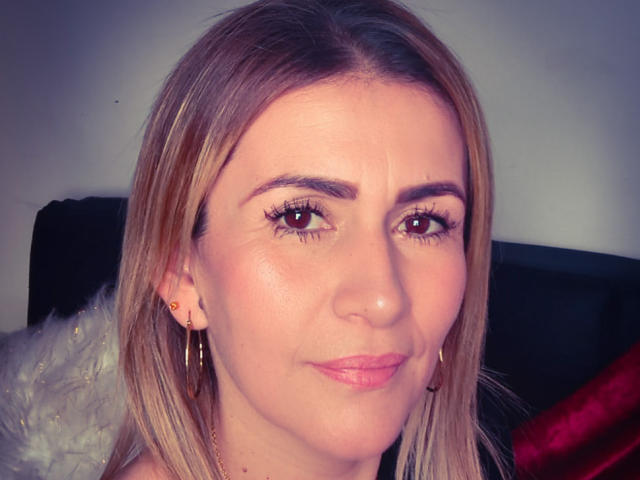 CamilleMullerr tem cabelo castanho claro, olhos castanhos e batom rosa. Ela está usando brincos dourados e tem um leve sorriso. Sua pele é clara, com um leve rubor..