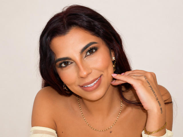 CarlaTorres has medium brown skin, dark wavy hair, and a cute smile. She's wearing gold earrings, a chain necklace, and a bracelet. Her hand's touching her cheek.