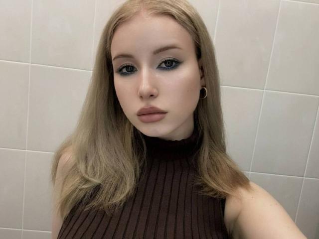 RefugioKerth, avec sa peau pâle, ses yeux bleus et ses cheveux blonds, portant un haut marron en maille côtelée sans manches, regarde directement l'appareil photo avec une expression neutre..